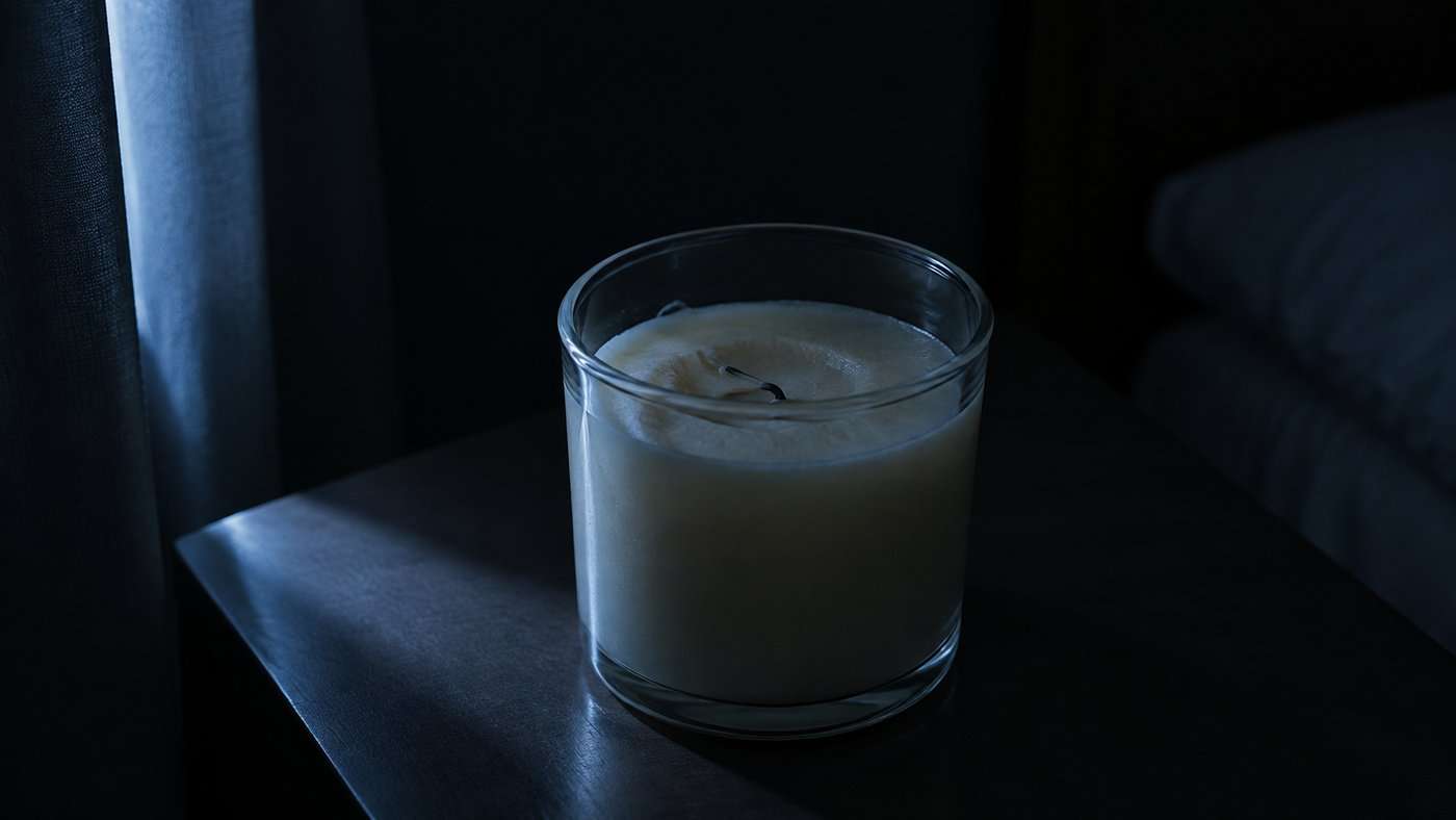 Close-up of a plain glass jar candle in a dark bedroom, its wax cooled and its spent wick lying flat and clearly unlit