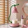 The pink bell lamp switched off on a desk with pens and a mirror, blinds striping the wall, a cute candle warmer