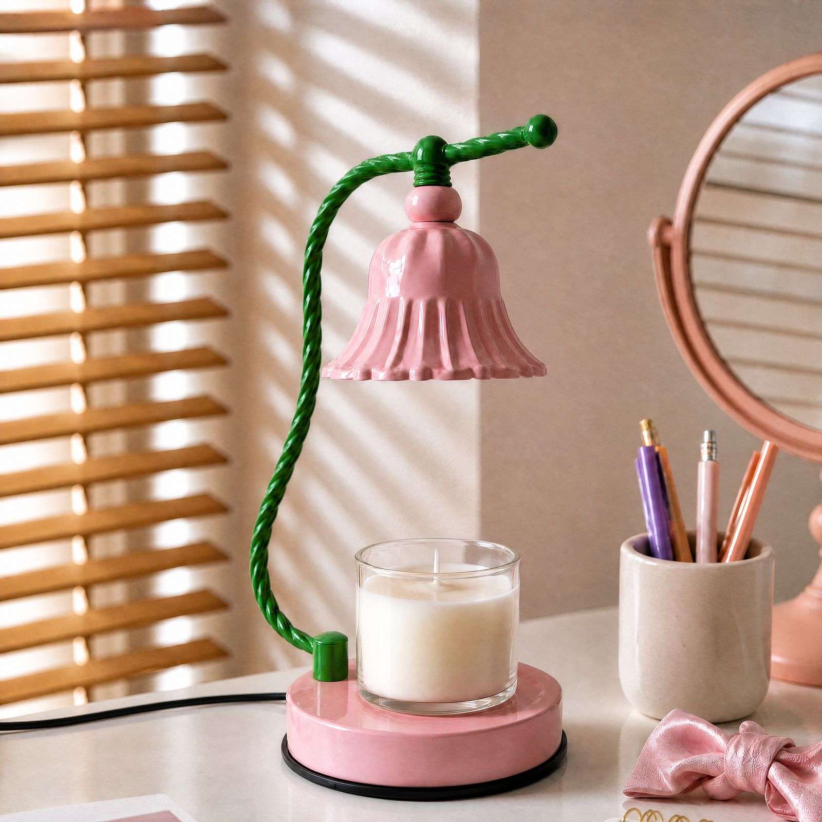 The pink bell lamp switched off on a desk with pens and a mirror, blinds striping the wall, a cute candle warmer
