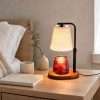Lit on a bedside table beside a book, a ceramic cup and eucalyptus in a vase, a candle lamp with shade in use