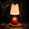 Dark still life: warm light through the linen-look shade onto a red jar, dried wheat behind, a rustic candle warmer