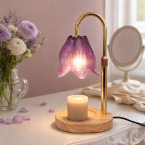 Purple glass tulip lamp lit over a white jar candle on a round wood base, on a dressing table with flowers and a mirror